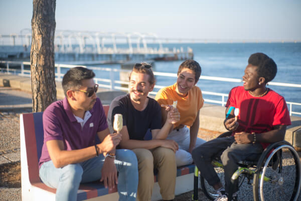 Trois personnes assises sur un banc public accompagnées d'une personne en chaise roulante, au bord de la mer en train de manger une glace et passer un bon moment